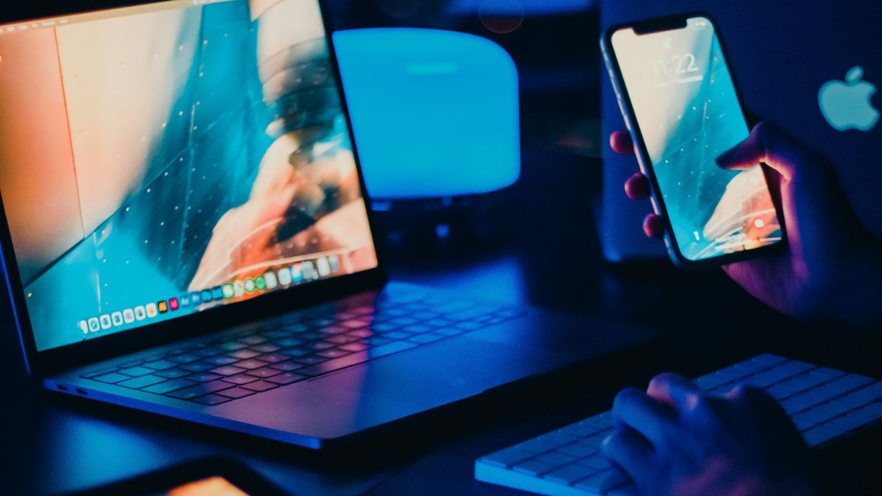 Notebook, tablet e celular iluminados em azul sobre uma mesa, em ambiente escuro de trabalho